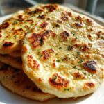 A stack of freshly made, golden homemade naan bread topped with melted butter and chopped parsley.