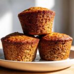 Close-up of three golden brown honey bran muffins stacked on a light plate, showing a moist, textured crumb.