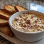 Close-up of a hot, cheesy reuben dip in a white bowl served with toasted rye bread slices.