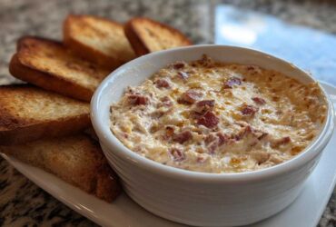 Close-up of a hot, cheesy reuben dip in a white bowl served with toasted rye bread slices.
