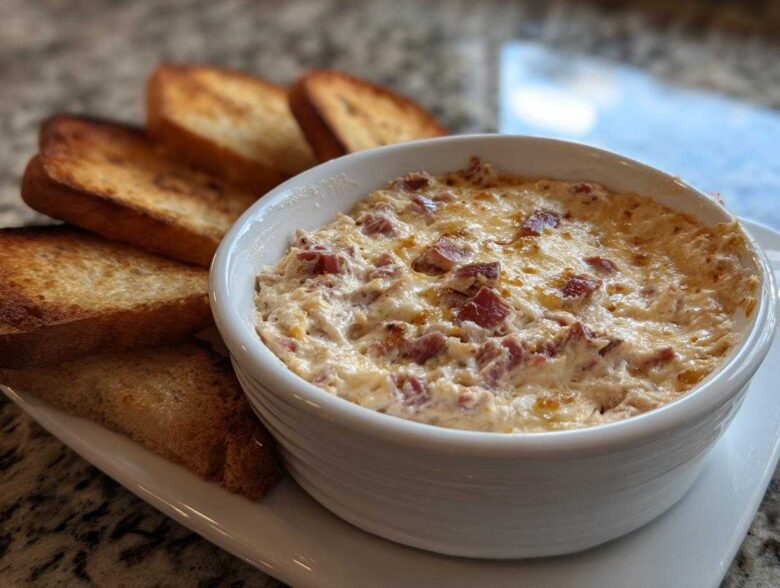 Close-up of a hot, cheesy reuben dip in a white bowl served with toasted rye bread slices.