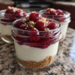 Three individual servings of Protein Cheesecake in small glass jars, layered with crust, creamy filling, and topped with cherry compote and crumble.