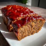 A close-up of a thick slice cut from a juicy Keto meatloaf topped with a shiny, dark red glaze.