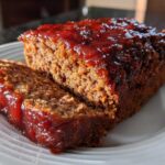 A thick slice cut from a juicy Keto meatloaf topped with a shiny, dark red glaze, served on a white plate.