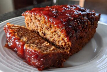 A thick slice cut from a juicy Keto meatloaf topped with a shiny, dark red glaze, served on a white plate.