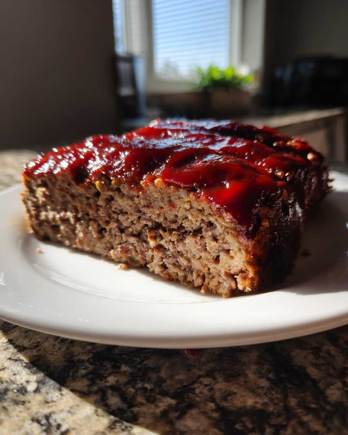 A thick slice of moist meatloaf topped with a shiny, dark red glaze, served on a white plate.