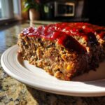 A thick slice of moist meatloaf topped with shiny red glaze, sitting on a plate.