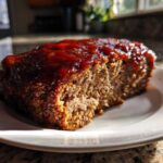 A thick slice of moist meatloaf topped with a shiny, thick red glaze, sitting on a white plate.