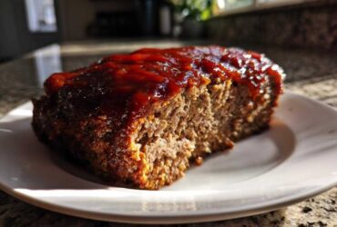 A thick slice of moist meatloaf topped with a shiny, thick red glaze, sitting on a white plate.