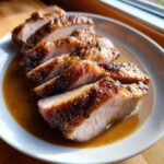 Close-up of perfectly cooked, sliced pork loin roast resting in a savory brown pan sauce on a white plate.