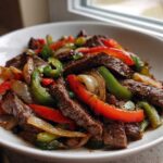 Close-up of sizzling steak fajitas featuring strips of seared beef, red and green bell peppers, and onions in a white bowl.