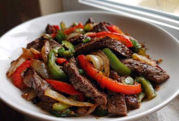 Close-up of sizzling steak fajitas featuring strips of seared beef, red and green bell peppers, and onions in a white bowl.