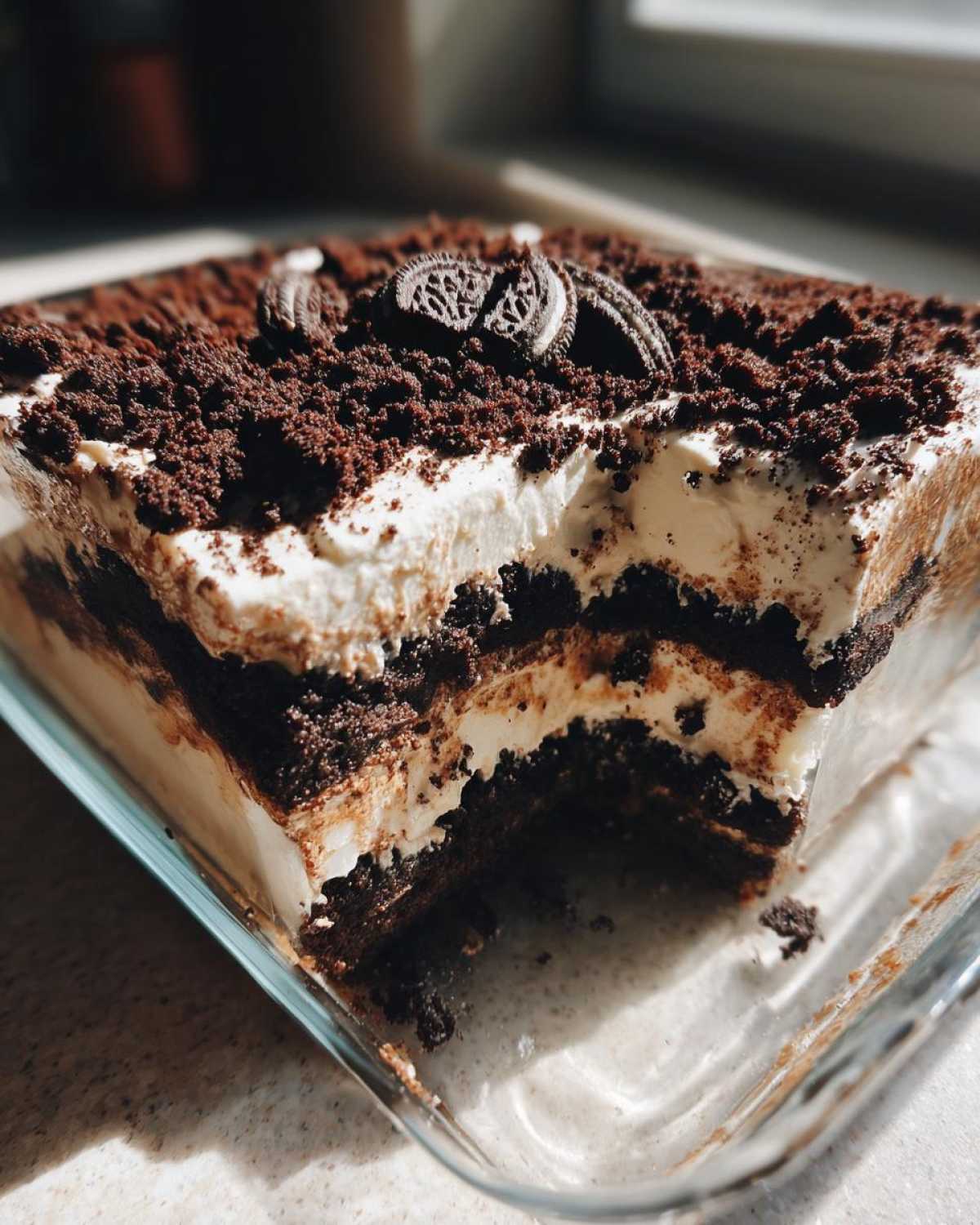 A square glass dish showing a slice taken out of a rich, layered dirt cake topped with crushed cookies and whole Oreos.