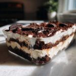 A clear glass dish filled with layered oreo dirt cake, showing alternating layers of cream and crushed chocolate cookies.