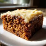 Close-up of a moist, dark slice of applesauce cake topped with thick cream cheese frosting on a white plate.