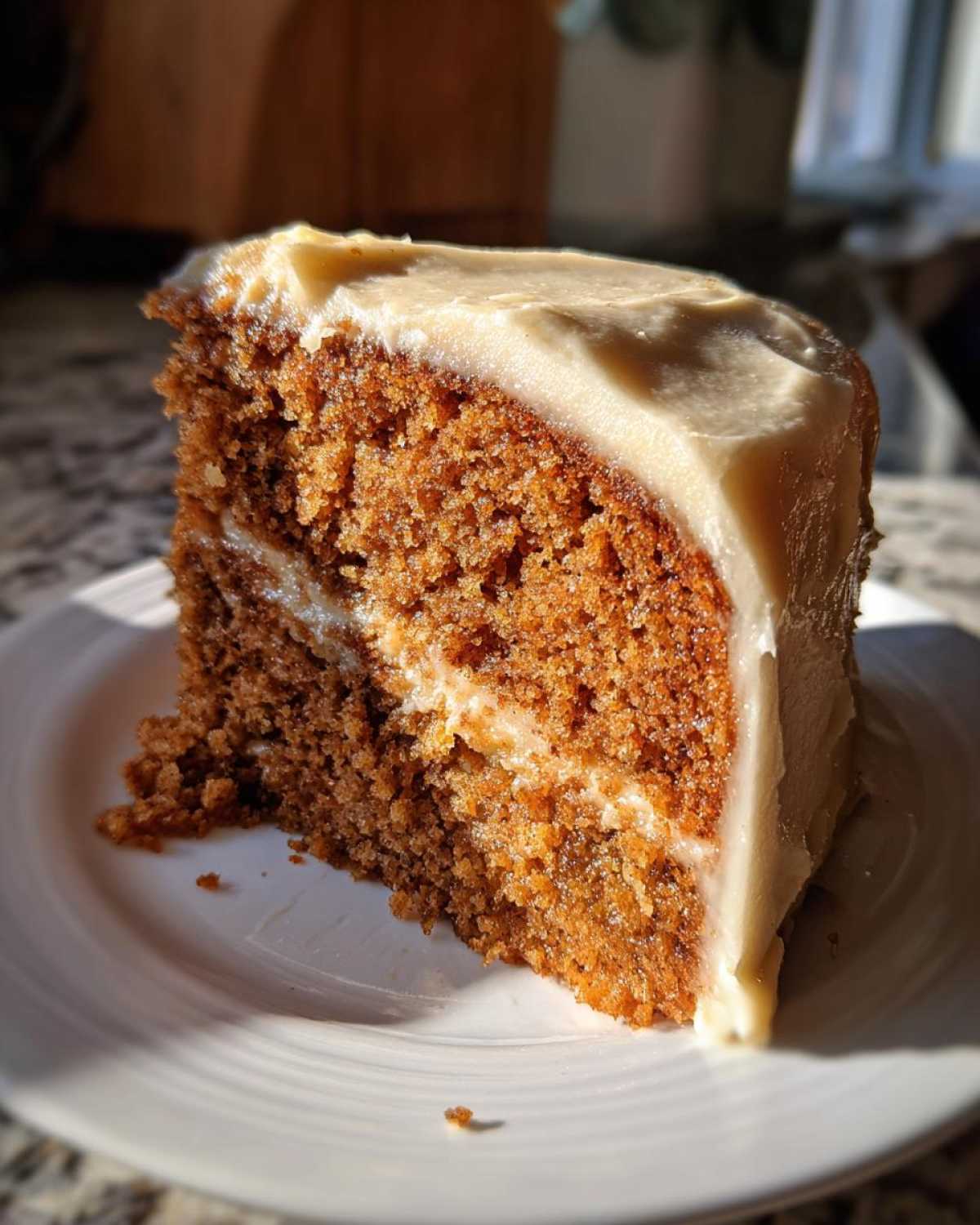 Close-up of a moist slice of applesauce cake with thick, creamy frosting, highlighted by sunlight.