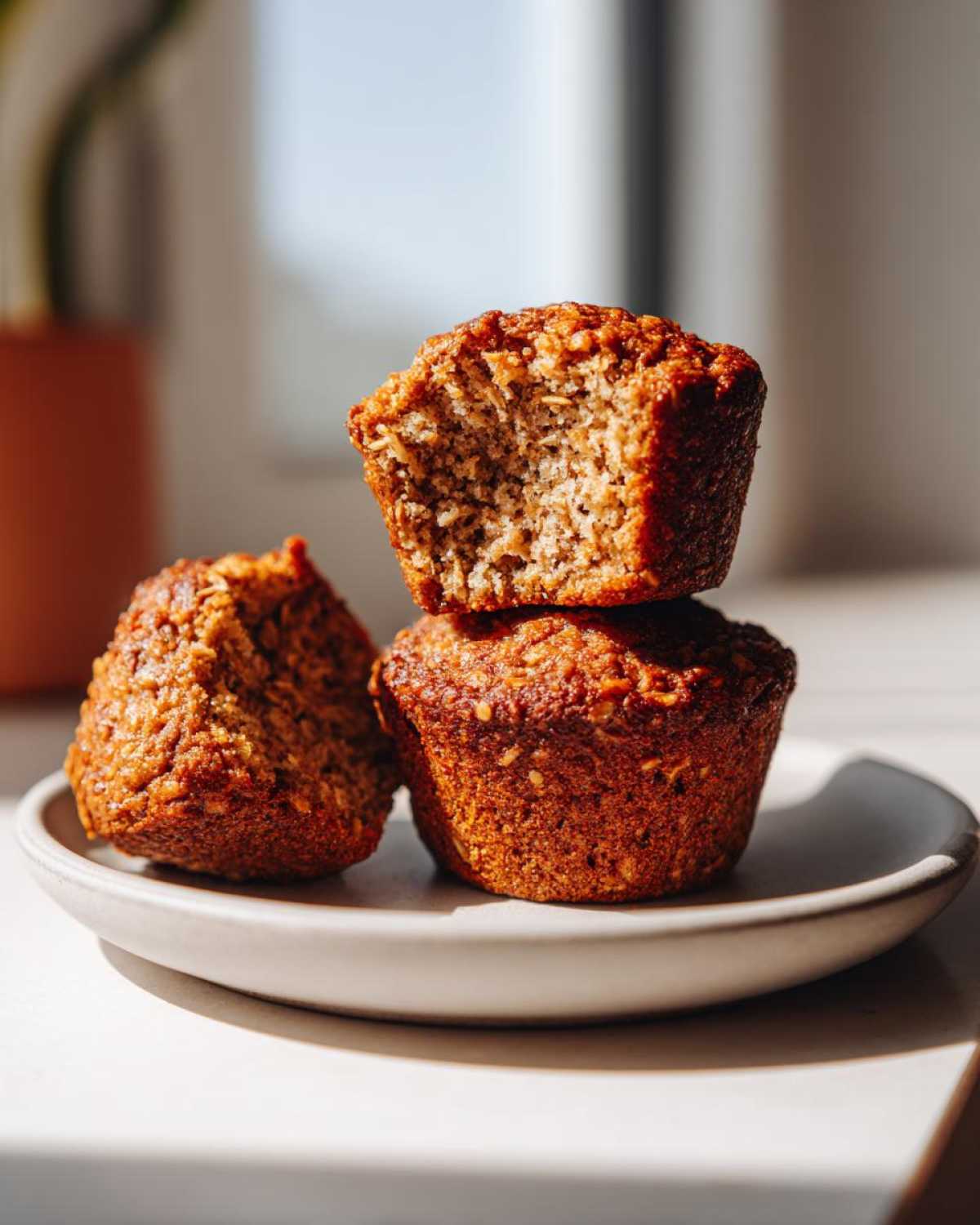 Three golden-brown bran muffins stacked on a small plate, one on top has a bite taken out showing the moist interior.