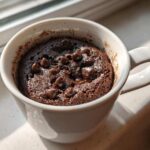 Close-up of a moist chocolate chip mug cake, baked in a white mug, sitting by a sunny window.