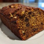 A close-up of a moist, dark brown date nut bread loaf, showing raisins and walnuts baked inside, resting on a white plate.