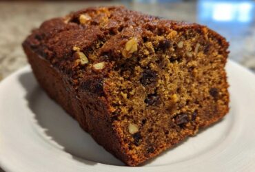 A close-up of a moist, dark brown date nut bread loaf, showing raisins and walnuts baked inside, resting on a white plate.