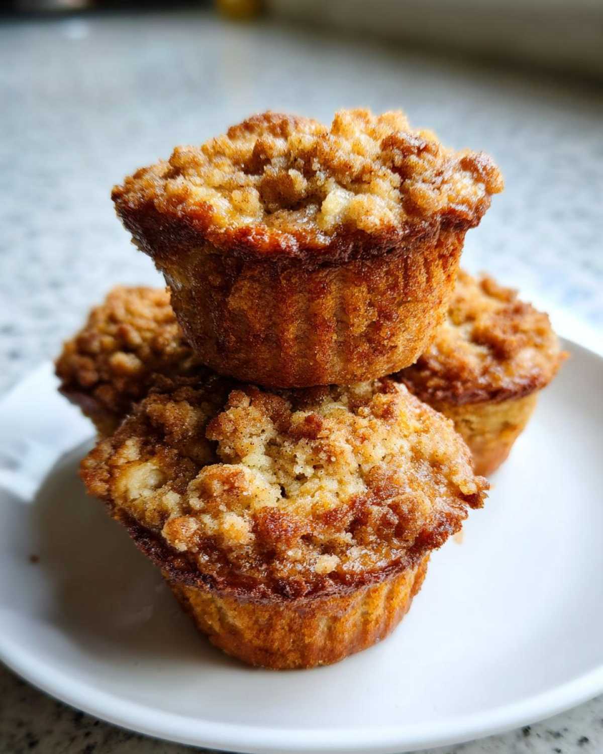 Three golden brown oatmeal muffins stacked on a white plate, featuring a thick, sugary crumb topping.