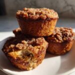 A stack of three golden brown oatmeal muffins with a sugary crumb topping, one muffin is broken open slightly.