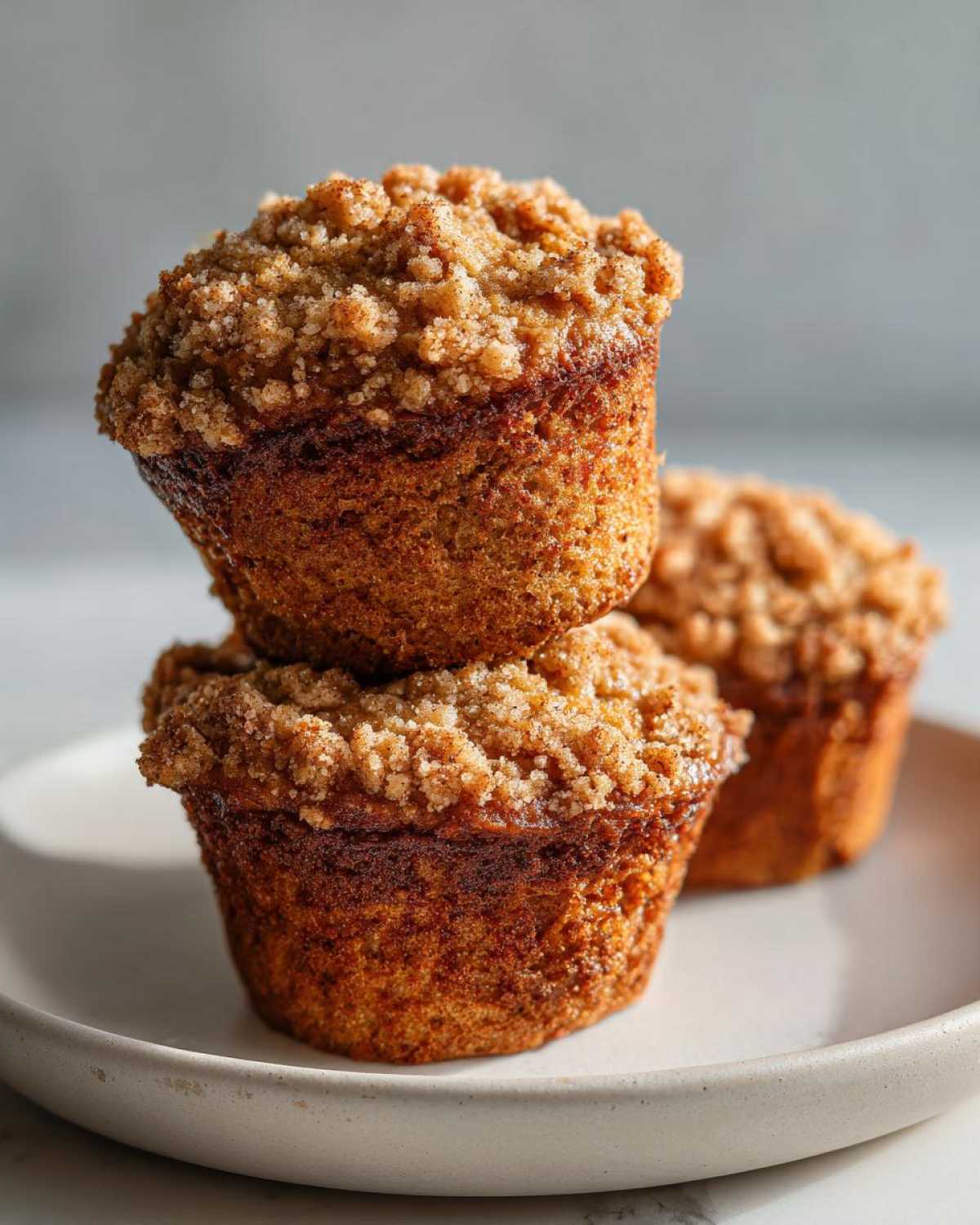 A stack of two moist oatmeal muffins topped with a generous cinnamon streusel topping, with another muffin blurred in the background.