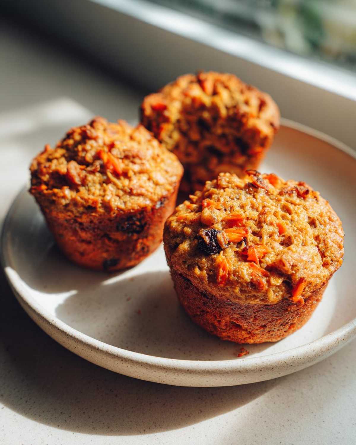 Three freshly baked morning glory muffins with visible carrots and raisins sitting on a light-colored ceramic plate near a window.