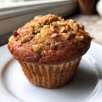Close-up of a golden brown banana nut muffin with a crumbly top sprinkled with chopped walnuts, resting on a white plate.