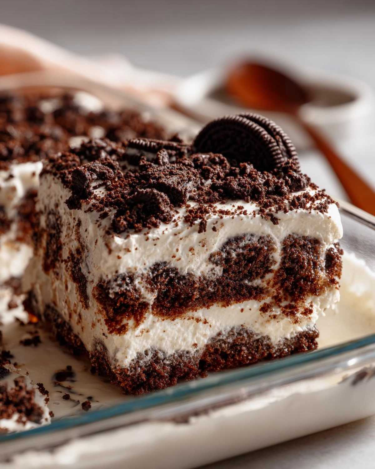 A close-up of a rich slice of oreo dirt cake featuring layers of chocolate cookie crumbs and white cream, topped with more crumbs and a whole Oreo.