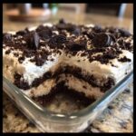 Close-up of a square glass dish of layered dirt cake, showing a missing slice revealing chocolate cookie layers and creamy filling, topped with whipped cream and Oreos.