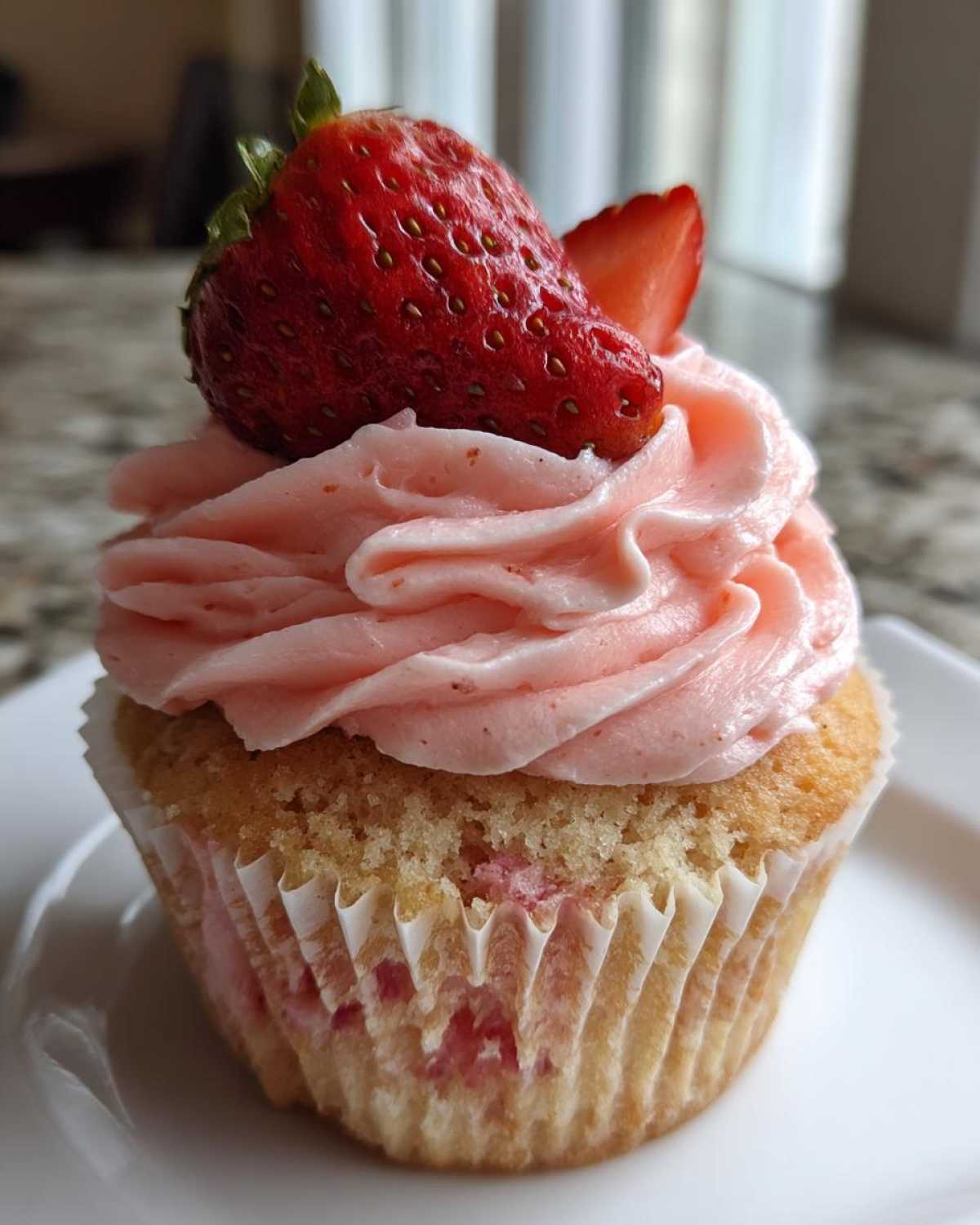 Close-up of a moist strawberry cupcake topped with pink strawberry frosting and a fresh whole strawberry.