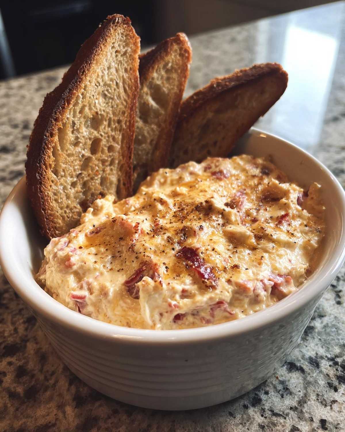 Creamy reuben dip served warm in a white bowl, topped with pepper, alongside toasted baguette slices for dipping.