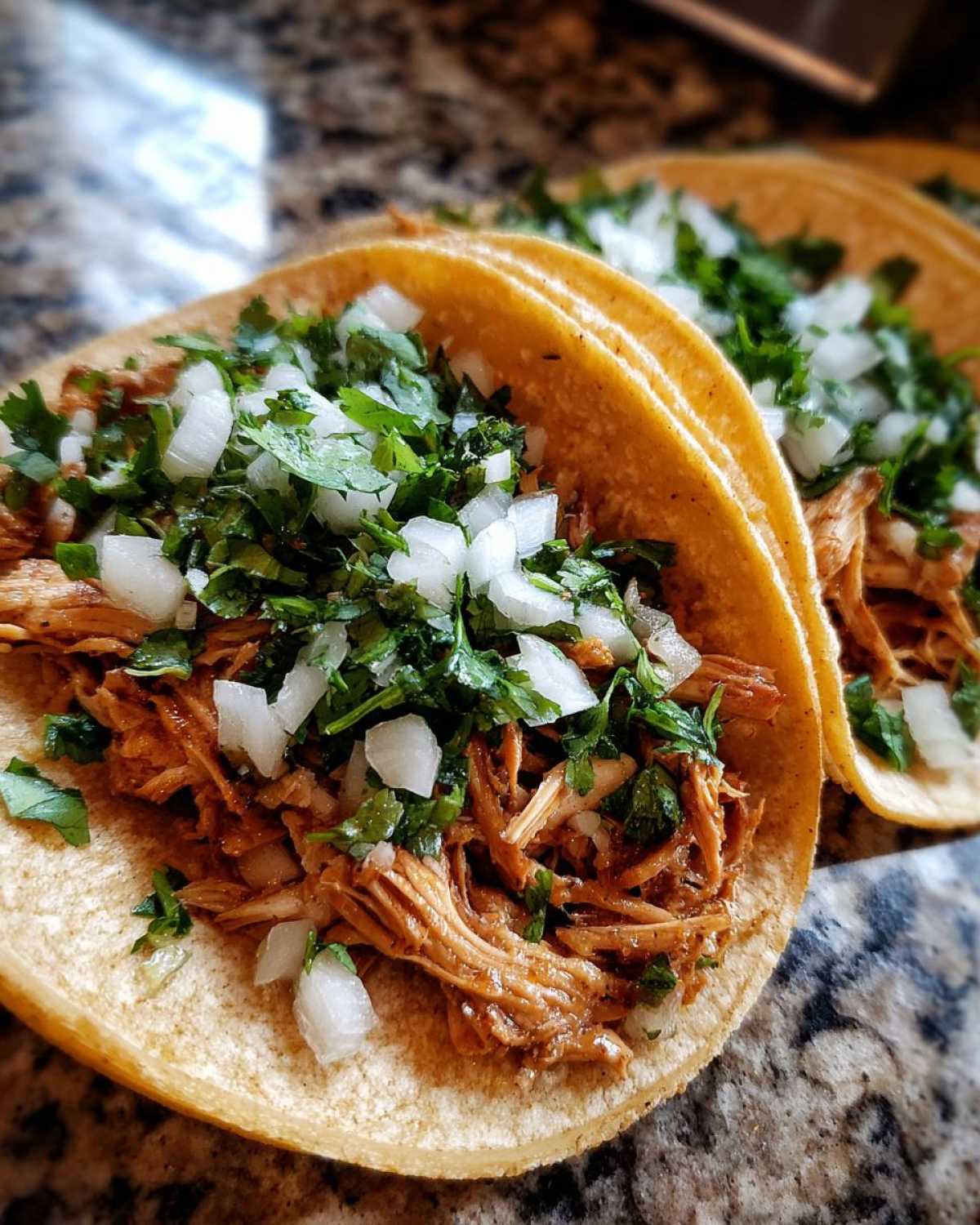 Close-up of two corn tortilla chicken tacos filled with shredded chicken and topped with fresh cilantro and diced white onion.