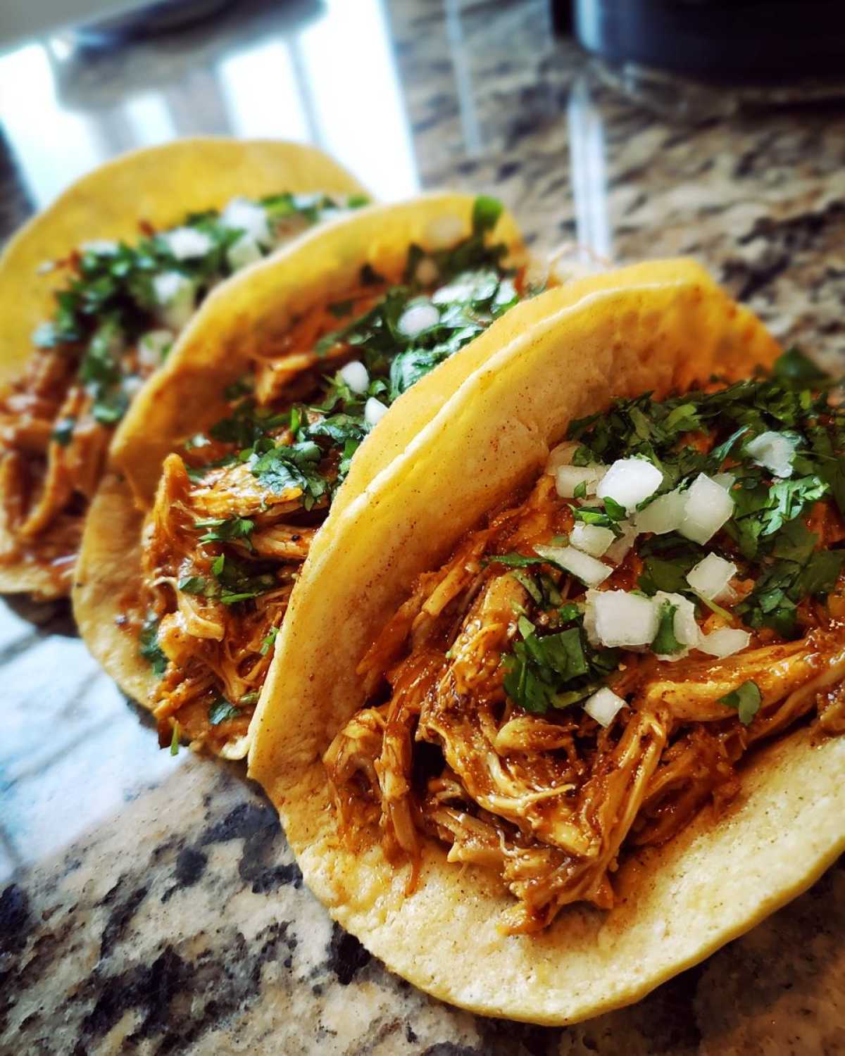 Three soft corn tortillas filled with saucy shredded chicken tacos, topped with diced onion and cilantro.
