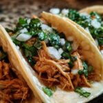 Close-up of three soft corn tortillas filled with shredded chicken tacos, topped with diced white onion and cilantro.