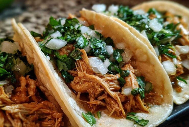 Close-up of three soft corn tortillas filled with shredded chicken tacos, topped with diced white onion and cilantro.