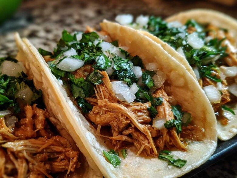 Close-up of three soft corn tortillas filled with shredded chicken tacos, topped with diced white onion and cilantro.