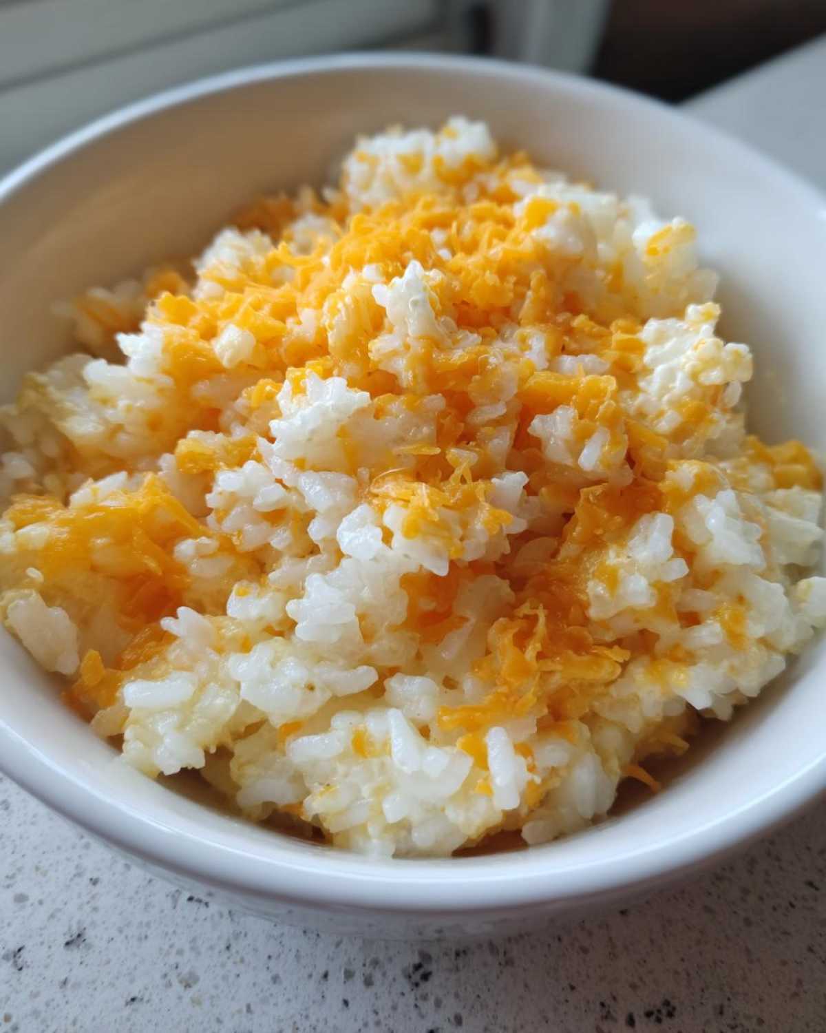 Close-up of a white bowl filled with fluffy white rice topped generously with melted shredded cheddar cheese, creating delicious cheesy rice.