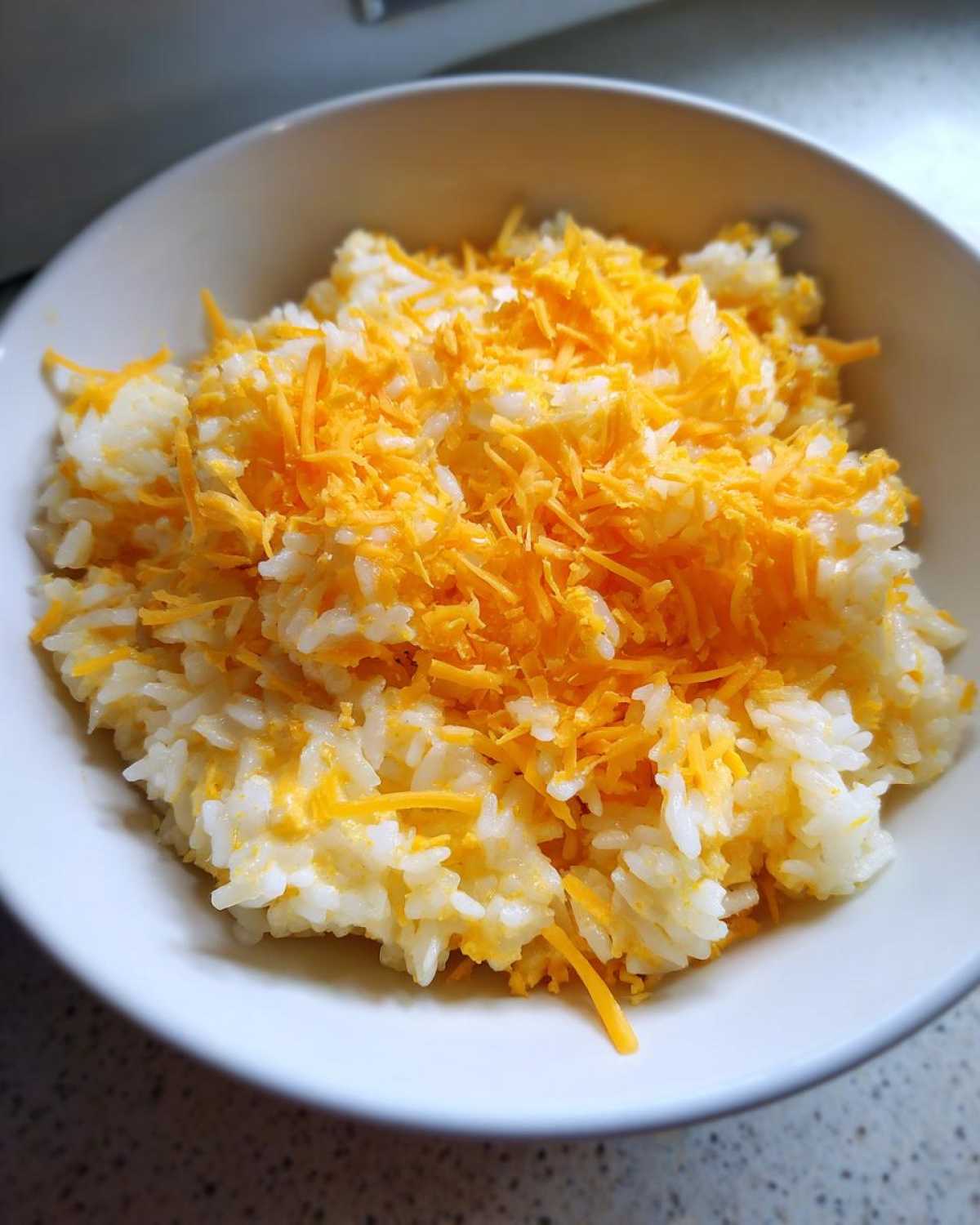 Close-up of a white bowl filled with fluffy rice topped generously with shredded cheddar cheese for cheesy rice.