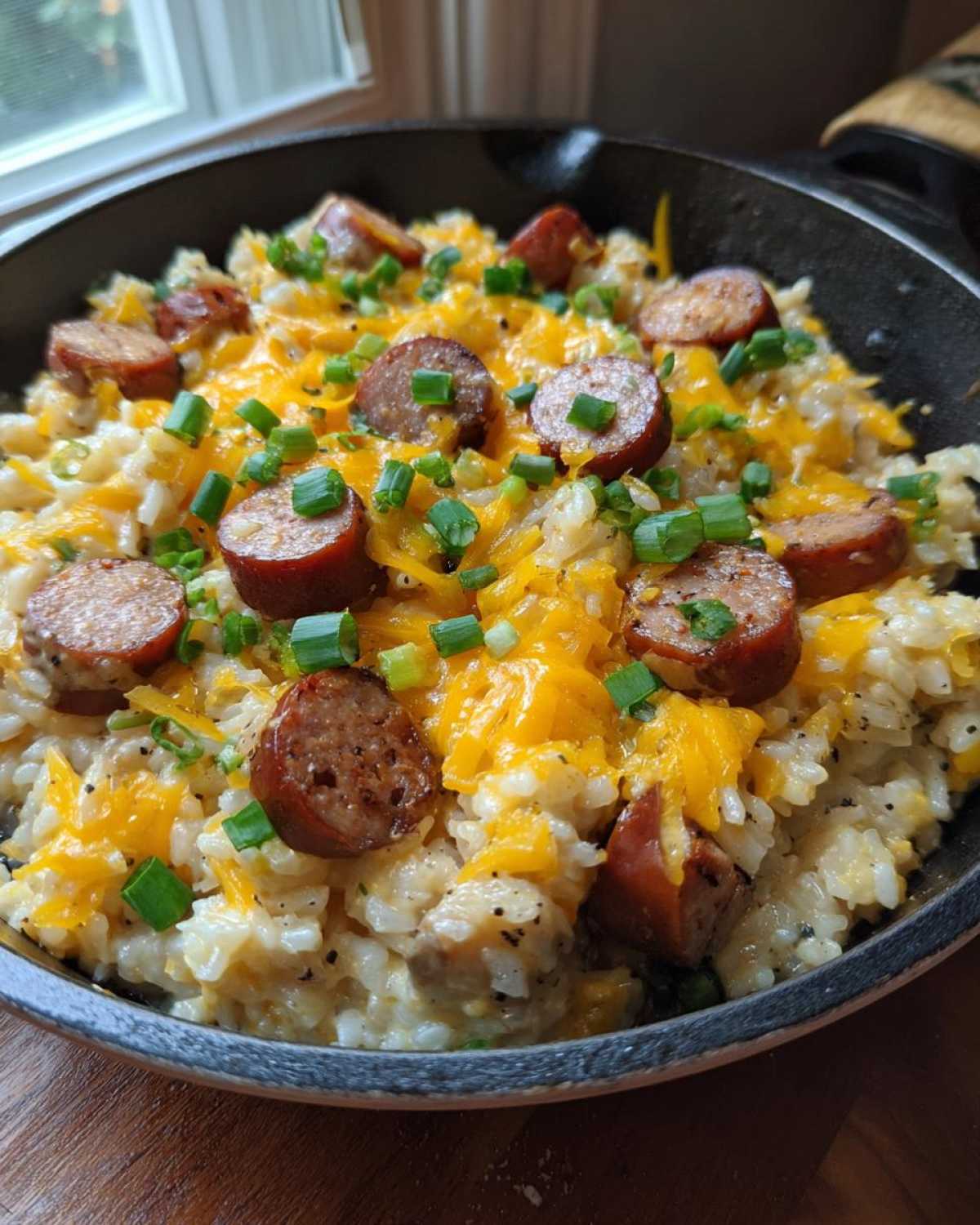 Close-up of cheesy sausage and rice cooked in a black skillet, topped with green onions.