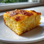 A square slice of moist cornbread casserole with visible corn kernels, served on a white plate.