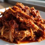 Close-up of a mound of saucy, shredded Slow Cooker Pulled Pork glistening under natural light on a white plate.