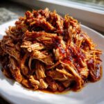 A close-up of a generous pile of saucy, shredded Slow Cooker Pulled Pork on a white plate.