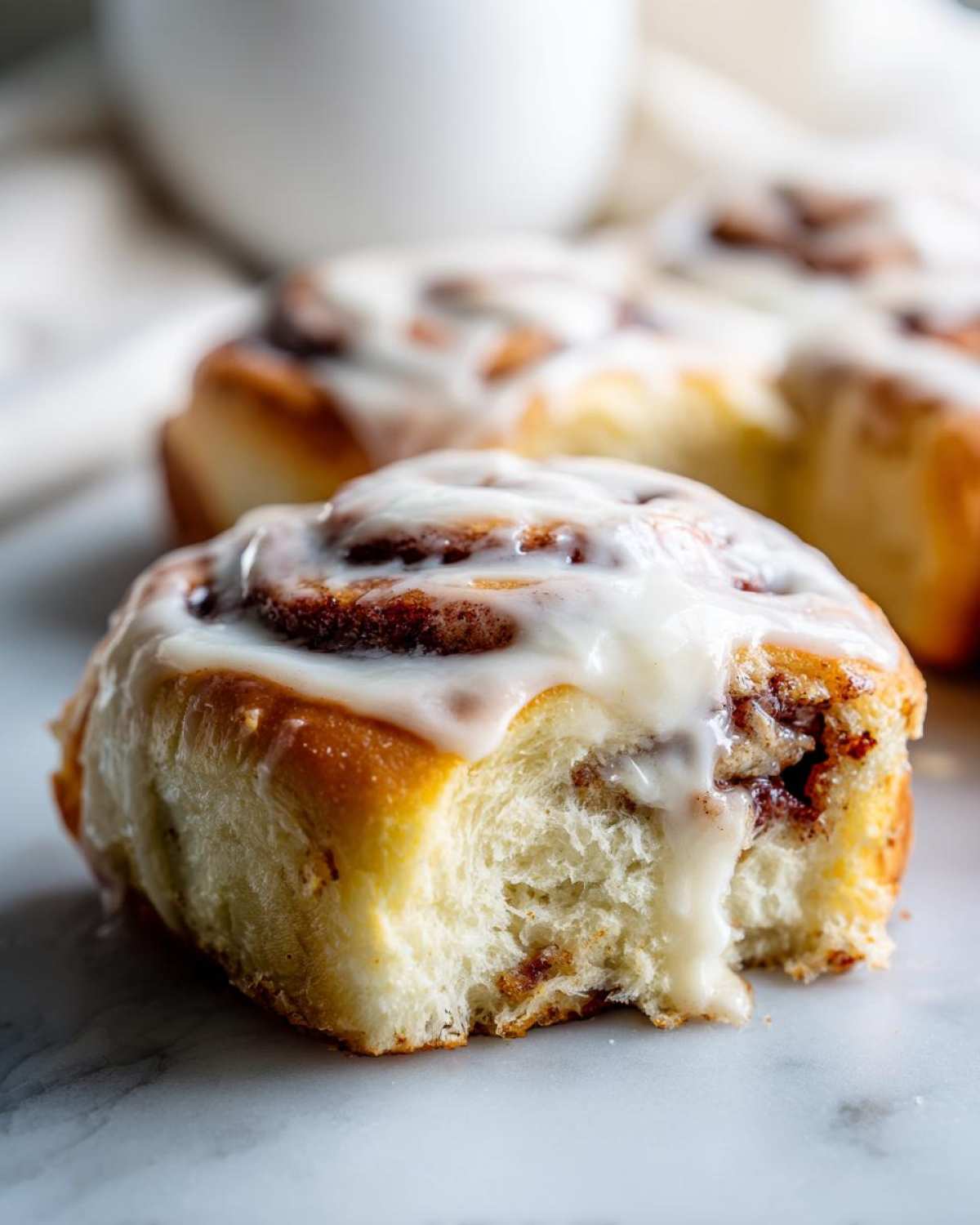 A close-up of a soft, fluffy cinnamon roll with a bite taken out, generously topped with dripping white icing.
