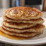 A close-up of a tall stack of five golden brown oatmeal pancakes drizzled with syrup on a white plate.