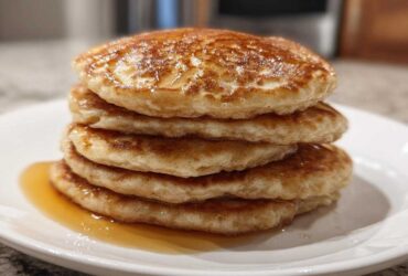 A close-up of a tall stack of five golden brown oatmeal pancakes drizzled with syrup on a white plate.