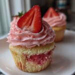 A close-up of a delicious strawberry cupcake with pink frosting and a fresh strawberry slice on top.