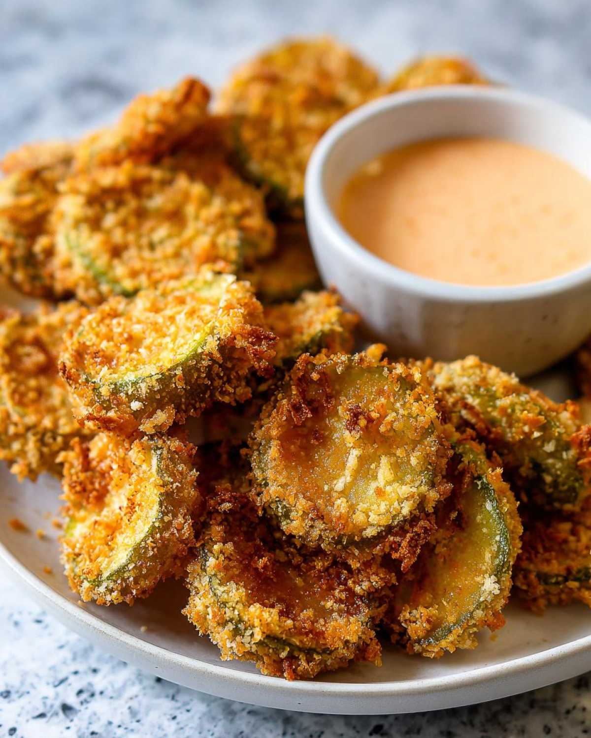 Close-up of crispy, golden brown Fried Pickles piled on a plate next to a small bowl of creamy dipping sauce.