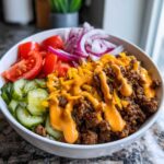 A close-up of a hearty cheeseburger bowl featuring seasoned ground beef, shredded cheese, special sauce, pickles, tomatoes, and red onion.